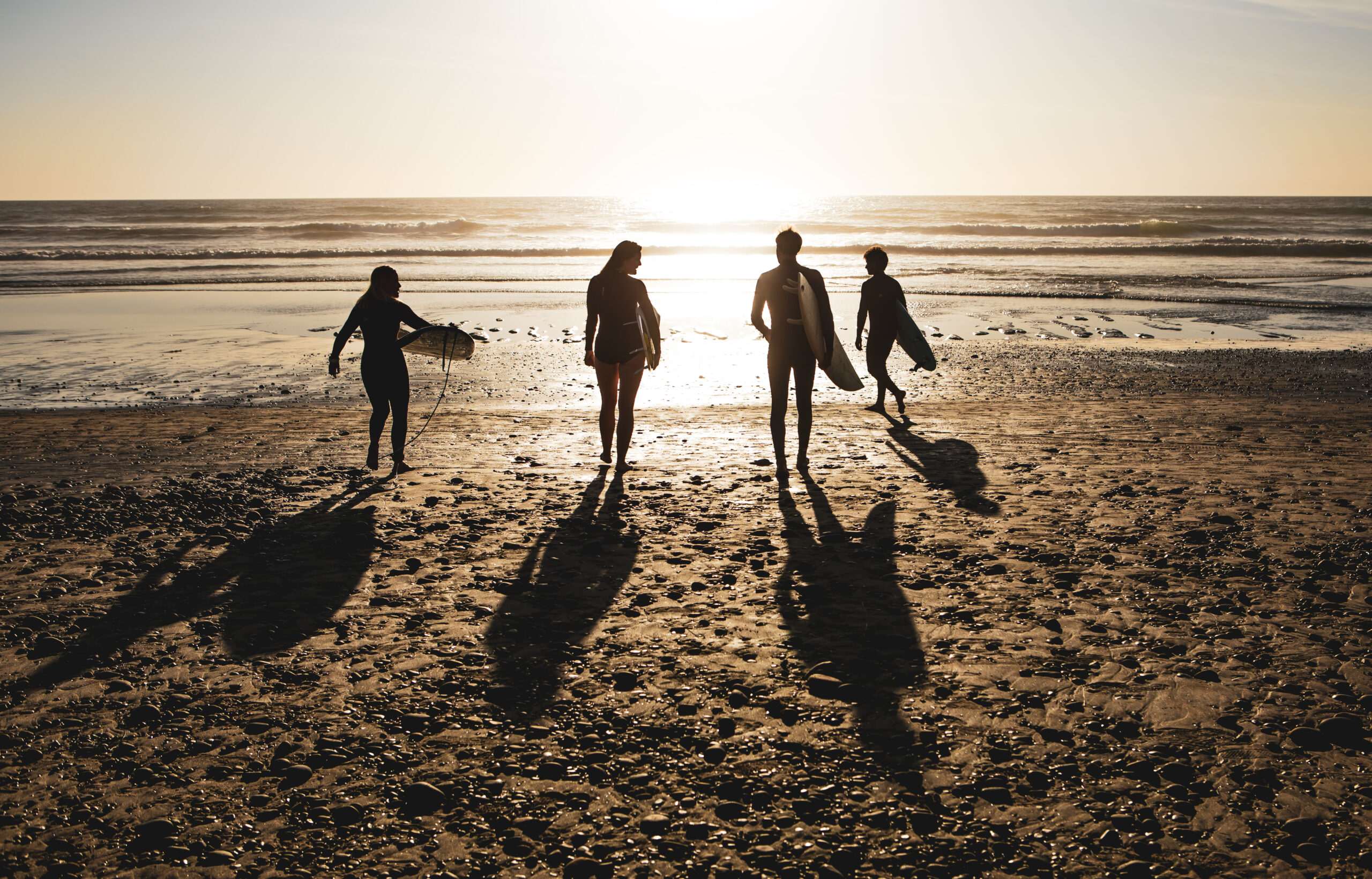 Four people walk on a sandy beach towards the ocean at sunset, carrying surfboards. Their long shadows stretch out behind them on the beach, creating an active scene perfect for an evening surf before unwinding with a glass of wine.