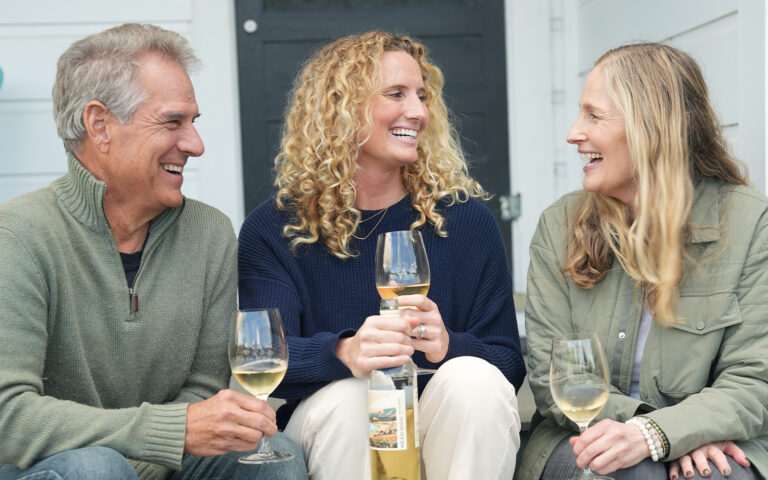 Bill Price, Kaleigh Gilchrist, and Prema Kerollis laughing on a porch drinking wine