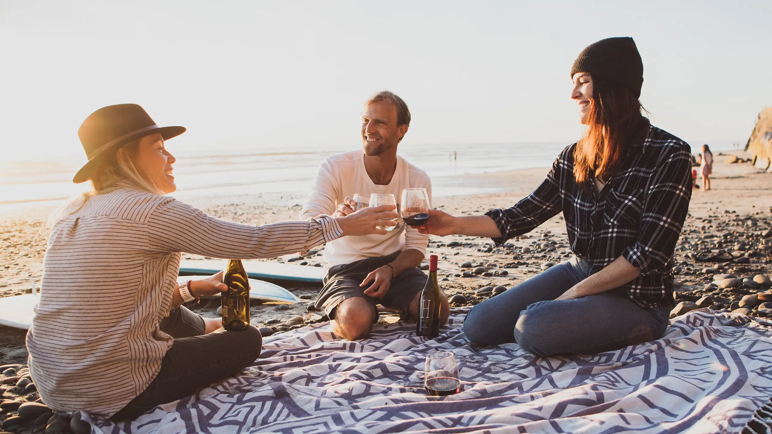 Three people sitting on a blanket at a beach, clinking glasses and smiling. The sun is setting as they relax in the great outdoors, with a bottle of wine and other items around them.