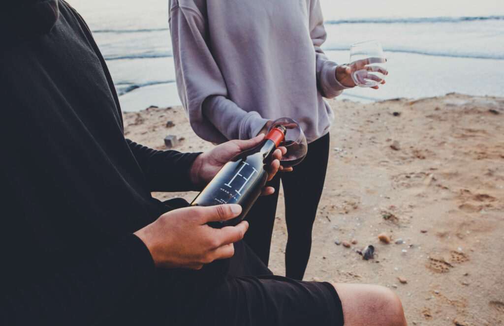 Two people by the beach, one holding a wine bottle and the other a wine glass, as the surf of Sonoma's coastline creates an enchanting backdrop.