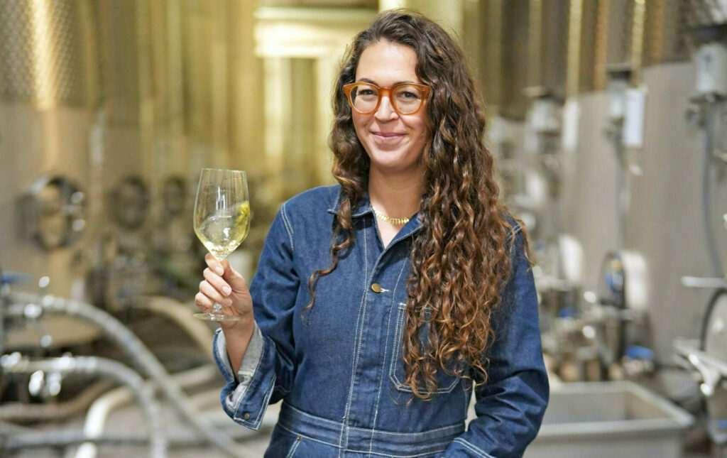 A person with long curly hair and glasses holds a glass of white wine while standing outdoors in a Sonoma winery, wearing a denim jacket. Stainless steel wine tanks are visible in the background.