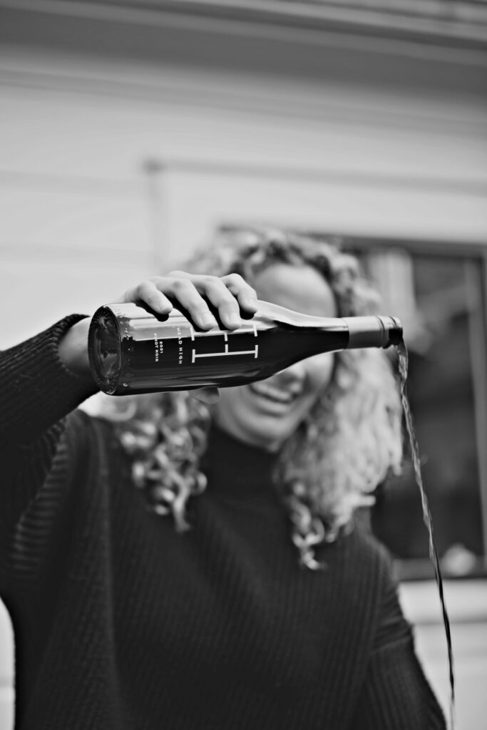 A person with curly hair wearing a dark sweater is tilting a wine bottle, pouring its contents out, with a building in the background.