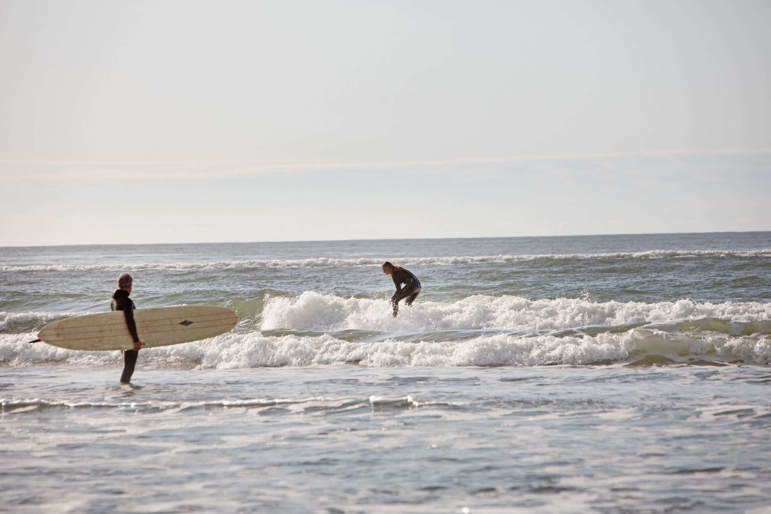 Two surfers at the beach, one standing in shallow water holding a surfboard and the other riding a wave.