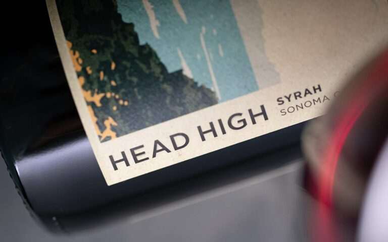 Close-up of a wine bottle labeled "Head High Syrah Sonoma Cou." A wine glass with red wine is partially visible in the foreground.